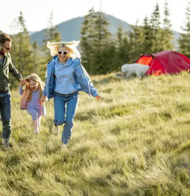 A joyful family dashes through a vibrant field, with a tent set up in the background under a clear blue sky.
